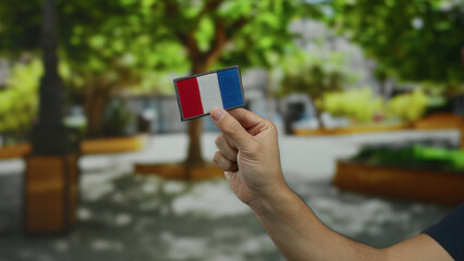 Man holding an embroidered french flag patch in a park, showcasing national pride amidst lush...