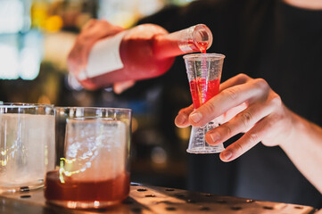 Bartender pouring red liquor into jigger at bar counter, with two glasses of ice in foreground....