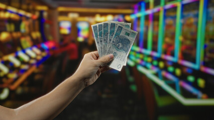 Man holding polish zloty banknotes inside a casino with slot machines in the background, creating a...