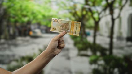 Man holding norwegian krone banknote outdoors on a city street, showcasing currency in a vibrant urban setting with blurred background trees and buildings.