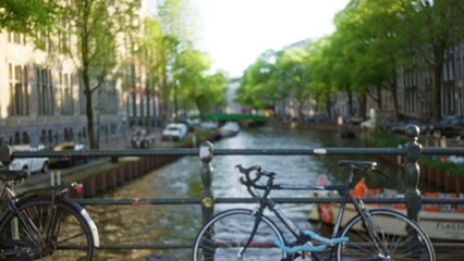 Blurred view of amsterdam canal with bicycles in foreground and defocused trees and buildings under bright sunlight creating a serene urban scene.