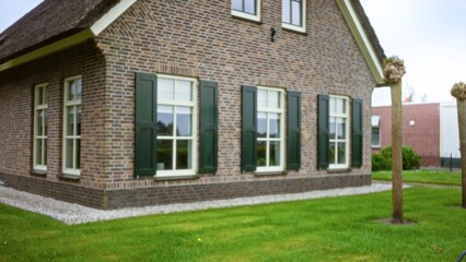 Blurred image of a traditional brick house with green shutters and manicured lawn, showcasing rustic architecture in a residential neighborhood with defocused trees in the forefront.