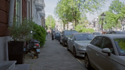 Blurred city street scene in a european town with parked cars, leafy trees, and people walking on a sunny day.