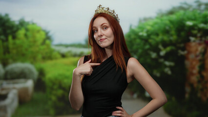 Woman wearing a crown stands confidently in a lush outdoor park setting, showcasing red hair and a...