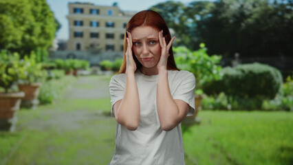 Woman in white shirt with headache stands in a sunlit park with green scenery and historic building...