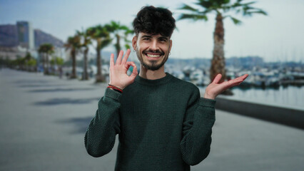 Smiling young man in sweater pointing at palm on a seaside promenade with palm trees and marina...