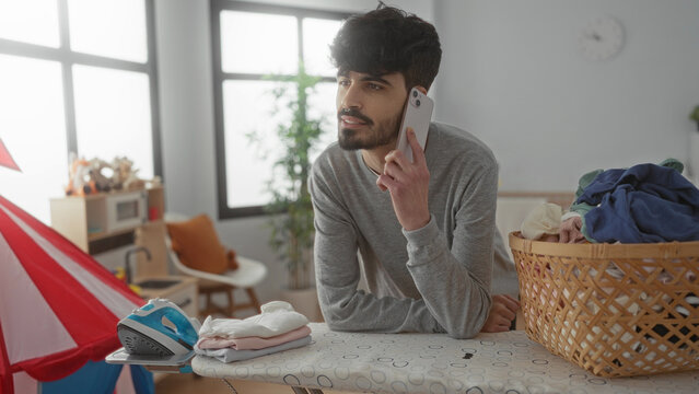Young man talking on phone while ironing in a bright laundry room with a basket of clothes and an ironing board, showcasing an attractive and casual home scene.