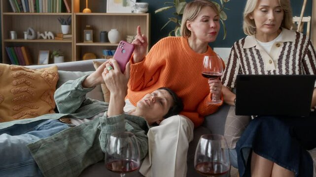 Group of three mature female friends relaxing together in living room, sipping wine, chatting and choosing good in online stores via laptop and smartphone
