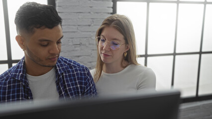 Man and woman collaborating in an office setting with natural lighting filtering through large...