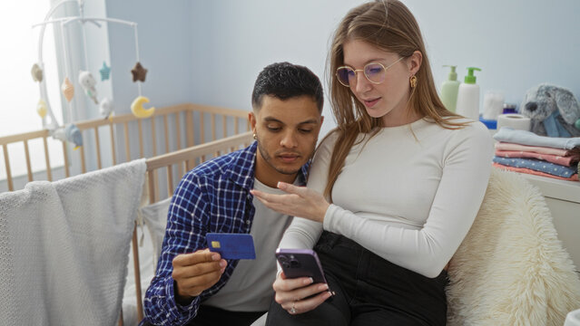 Couple shopping online in bedroom with credit card while woman holds phone in cozy setting beside baby cradle and man assists.