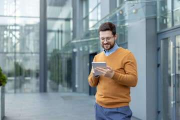 Smiling young male office worker standing outside the building and using a tablet in his hands