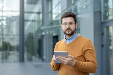 Close-up portrait of a young male office worker standing near a building, holding a tablet and...