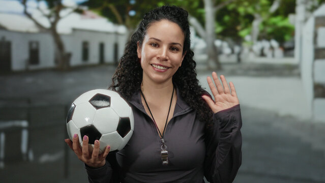 Young hispanic woman smiling outdoors holding soccer ball on the street with trees and buildings in the background