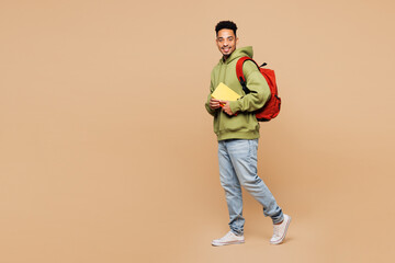 Full body side view young smart boy student he wear green casual clothes backpack bag hold books walking going isolated on plain pastel light beige background. High school university college concept.