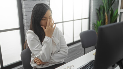 Woman in office appearing tired with closed eyes and hand on face in a modern workplace setting, featuring natural light through large windows and indoor plants.