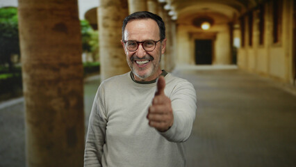 Mature hispanic man smiling in old university corridor offering handshake showcasing friendly...