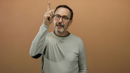 Man posing thoughtfully against brown background in casual outfit and glasses appearing inquisitive and engaged.