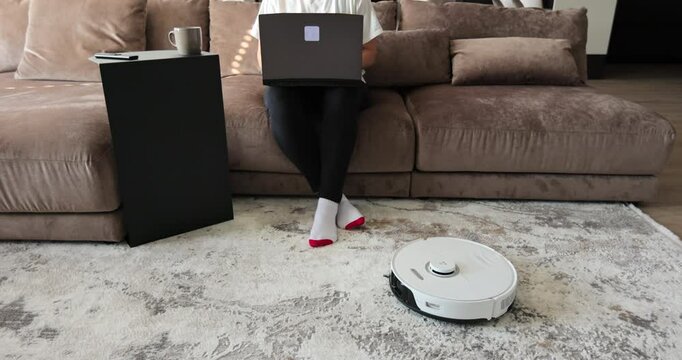 Young freelancer business woman using laptop, robot cleaner cleaning carpet. Remote work.