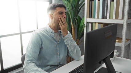 Young man yawning at desk in a modern office with a computer and bookshelf, suggesting workplace...