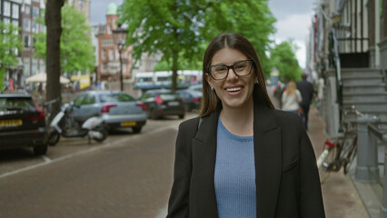 Woman smiling on amsterdam canal street, brunette adult in glasses enjoys a sunny day featuring urban architecture and parked cars along historic canals in the netherlands.