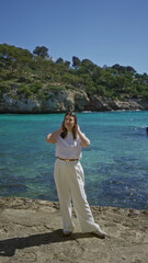 Woman smiling in white outfit enjoys sunny day by the turquoise waters and rocky cliffs at santanyi beach in mallorca with lush greenery surrounding the mediterranean landscape.
