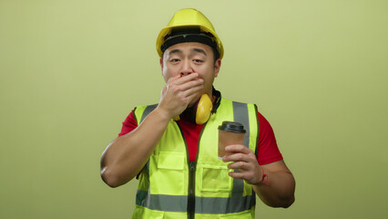 Tired asian construction worker yawning holding coffee on yellow background wearing safety gear and...