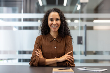 A smiling businesswoman in a modern office setting. She has curly hair and is wearing a brown...