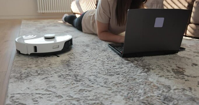 Woman push start button on robot vacuum cleaner. Woman working with laptop. Automatic vacuum cleaner. Smart home and easy life concept.