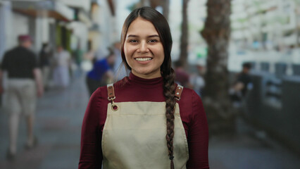 Hispanic woman waitress in apron smiling outdoors on city street with blurred background of people...