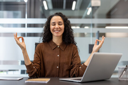 A smiling woman in a brown shirt meditates with her hands in a mudra at her desk in a modern office setting.