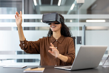A woman wears a vr headset and interacts with a virtual world, smiling at her workspace, with laptop and tablet.