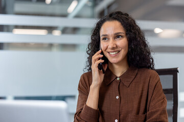 A smiling woman with curly hair is talking on the phone in a modern office setting, appearing...