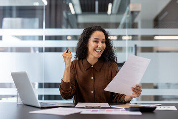 A joyful woman in an office celebrates a success, holding a document and smiling brightly. She is...