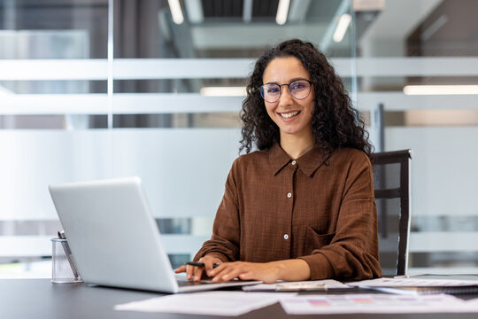 A smiling woman works on a laptop in a modern office environment, radiating professionalism and positivity.
