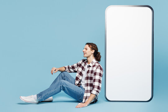 Full body side view young man he wear purple checkered shirt casual clothes sit near big huge blank screen mobile cell phone smartphone with area isolated on plain pastel light blue cyan background. - Powered by Adobe