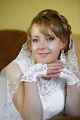 A woman in a white dress is sitting on a couch with her hands on her lap. She is wearing a lace...