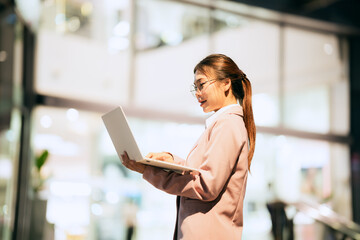 A young Asian businesswoman stands confidently outdoors in a modern urban setting, using a laptop....