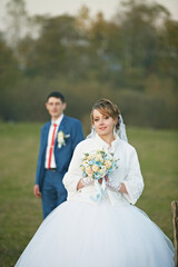 A bride and groom are standing in a field with a bouquet and a white jacket. The bride is holding a...