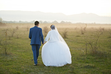 A bride and groom are walking through a field on their wedding day. The bride is wearing a long...