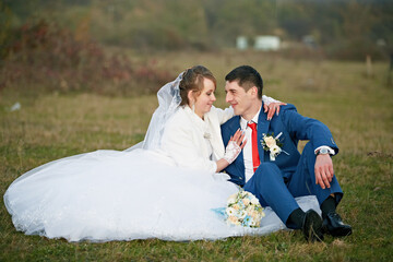 A bride and groom are sitting on the grass, the bride is wearing a white dress and the groom is...