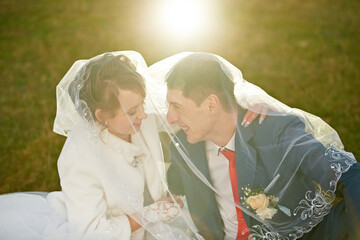 A bride and groom are sitting under a white veil, with the bride's face partially hidden. Scene is...