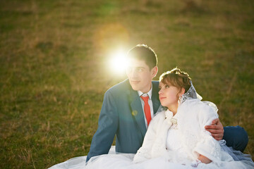 A bride and groom are sitting on the grass, enjoying the sun. The bride is wearing a white dress...