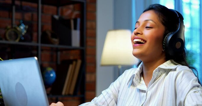 Indian businesswoman smiling attending video call on laptop using headphone at desk in modern cozy office, Asian young corporate executive expressing positivity while engaged in virtual meeting - Powered by Adobe