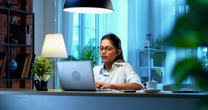 Indian businesswoman drafting email on laptop at desk in modern cozy office, Asian young female corporate executive typing an important message while thinking working remotely on wireless computer