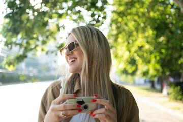 A young blonde woman hipster, smiling and photographing with a retro camera outdoors in the city...
