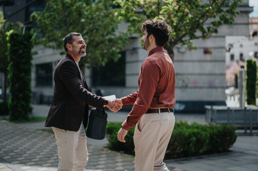 Two people exchange a friendly handshake in an urban street setting, surrounded by trees and modern...