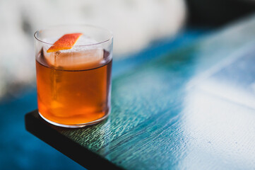 Elegant orange cocktail with ice cube and citrus garnish in a glass on wooden table, captured in moody lighting with blurred background.