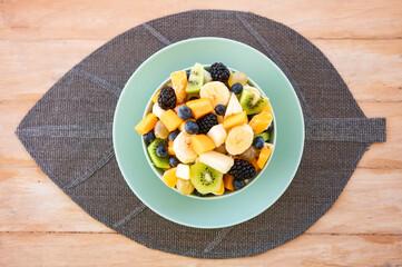 Delicious assortment of sliced fruits in a bowl for a fresh mixed fruit salad, served on a mint-green plate with a woven placemat over a rustic wooden table