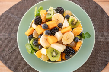 Delicious assortment of sliced fruits in a bowl for a fresh mixed fruit salad, served on a mint-green plate with a woven placemat over a rustic wooden table