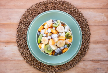 Two hands of a senior people holding a delicious assortment of sliced fruits in a bowl for a fresh mixed fruit salad, served on a mint-green plate with a woven placemat over a rustic wooden table
